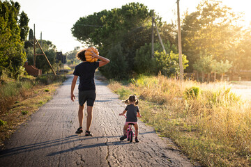 Little child girl, riding a bike, with her young father carrying a big halloween pumpkin on a...