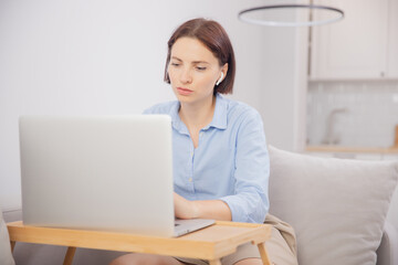 Lifestyle home office, young woman in headphones works laptop via Internet video call from sofa, light background