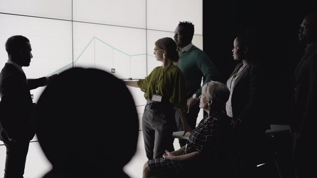 Diverse group of businesspeople going over charts
and graphs on a digital multiscreen wall during a
presentation in a dark office