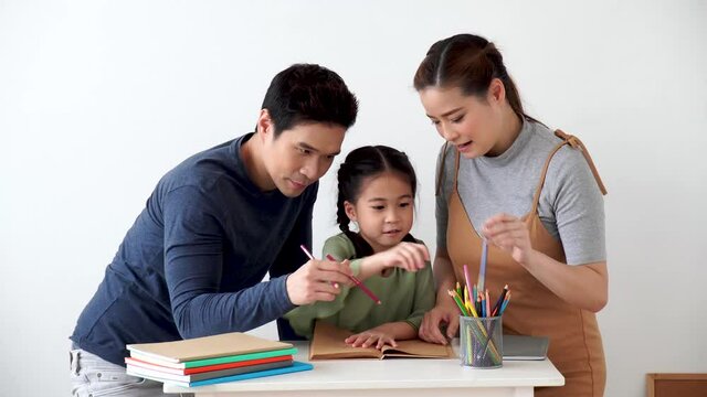 Asian young mother and father with little daughter sit at desk learning and writing in book with pencil making homework at home.Education concept.