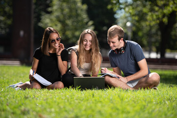 Three smiling college students learning and working together in a laptop while sitting on the ground of a park