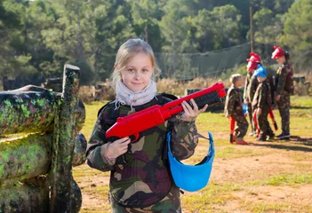 Tuinposter Portrait of cheery tween girl paintball player with marker gun ready for game outdoors © JackF