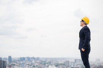 Young handsome asian civil engineer standing wearing helmet yellow hard hat in business suite on building urban cities background.