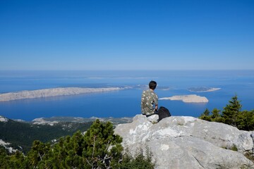 The beautiful Premuziceva Staza mountain path overlooking the Adriatic Sea and islands, Velebit National Park, Dinaric Mountains, Croatia. Lonely silhouette of young man sitting on rock in viewpoint.