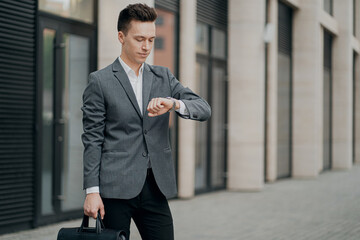 serious business man businessman, looking at the clock. fashionable barbershop hairstyle. thinking about a business meeting in the future. he holds the bag in his hands. background the city