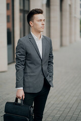 serious business man businessman, looking at the clock. fashionable barbershop hairstyle. thinking about a business meeting in the future. he holds the bag in his hands. background the city