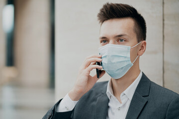 blue protective mask from the coronavirus 19 pandemic on the face. serious smart male student holding a new phone writes a friend a message. Grey business suit, dark hair