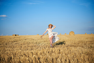Young blond woman, wearing white romantic dress and straw hat and dried grass bouquet, jumping running on straw field in summer. Female portrait on natural background. Environmental protection.