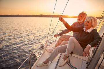 Enjoying luxury life. Beautiful happy senior couple in love relaxing on the side of sailboat or yacht deck floating in sea at sunset, looking at amazing evening view - Powered by Adobe