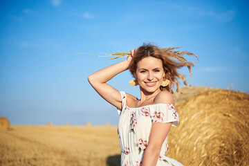 Fototapeta na wymiar Young blond woman, wearing long white romantic dress, holding straw hat and dried grass bouquet, posing in front of wheat bale on field in summer. Female portrait on natural background.