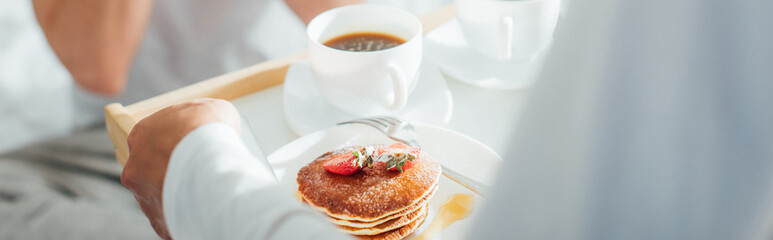 Panoramic crop of woman holding breakfast tray with tasty pancakes and coffee near man in bedroom