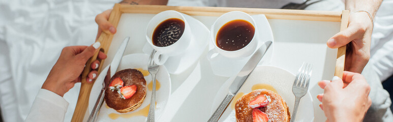 Horizontal concept of young woman holding breakfast tray with coffee and pancakes near man on bed