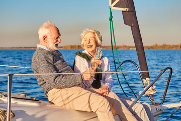 Fototapeta na wymiar Having fun. Happy beautiful senior family couple drinking wine or champagne and laughing while relaxing on a sailboat or yacht deck floating in a calm blue sea