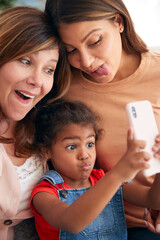 Multi-Generation Female Hispanic Family At Home Pulling Funny Faces For Selfie On Mobile Phone