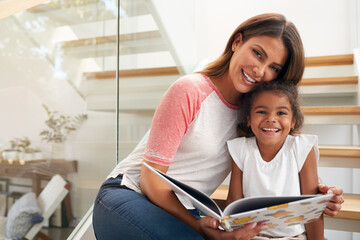 Portrait Of Hispanic Mother And Daughter Sitting On Staircase In Modern Home Reading Book Together