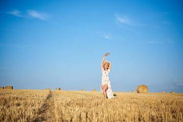 Young blond woman, wearing white romantic dress and straw hat and dried grass bouquet, walking on straw field in summer. Female portrait on natural background. Environmental protection.
