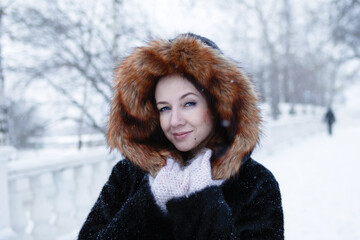 Portrait of young beautiful woman with blue eyes in black faux fur coat with red hood on a walk in...