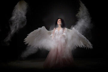 Girl with white wings posing in dark black studo during photoshoot with flour or dust and light....