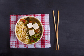 Japanese soup with tofu cheese, noodles, green beans, vegetables, herbs, bamboo chopsticks on a red napkin on a black background. Copy spaes. horizontally