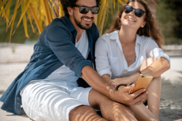 A beautiful young couple joyfully sits on white sand under a palm tree on the seashore under a green palm tree. Honeymoon Travel and Vacation on the Tropical Ocean.