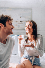 Young woman looking at boyfriend and sitting on bed with cup of coffee