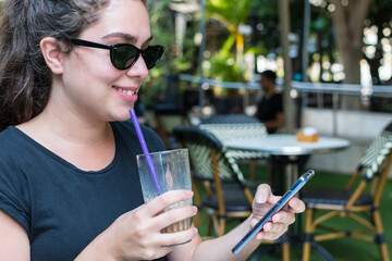 Young woman with sunglasses, drinks a fresh smoothie, sitting in an outdoor bar.