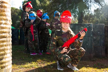 Tuinposter active girl paintball player in camouflage standing with gun before playing outdoors © JackF