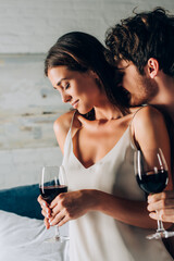 Selective focus of man kissing neck of girlfriend with glass of wine in bedroom