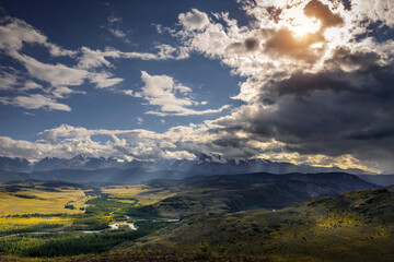 Fototapeta na wymiar Majestic panorama of mountain plain on the background of snow-covered ridge before thunderstorm. Sun's rays break through huge clouds and beautifully illuminate green steppe and winding river.