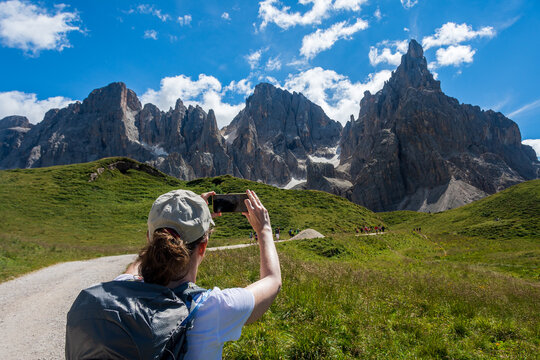 Young woman with a backpack and a hat stops on a mountain trail in the Italian Alps to take a picture with her phone to Pale di San Martino Dolomite mountain group. Val Venegia -  Trentino, Italy