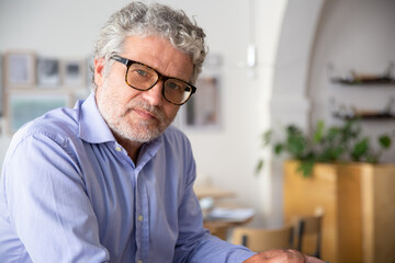 Serious pensive mature business man wearing shirt and glasses, sitting in office cafe, looking at camera. Medium shot, copy space. Business portrait concept