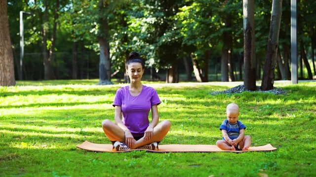 Young woman and baby sitting on yoga mat in park, female meditating and doing exercises, child looking around. Fit mother and son training together outside. Concept of fitness