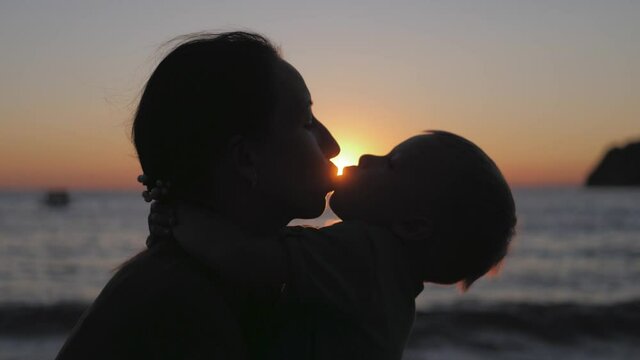 A mother and little son kiss on the beach and sea outdoors at sunset. Concept of happy vacation and friendly family.