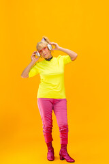 Listening to music. Senior woman in ultra trendy attire isolated on bright orange background. Looks stylish and fashionable, forever young. Caucasian model in sunglasses, bright attire and sneakers