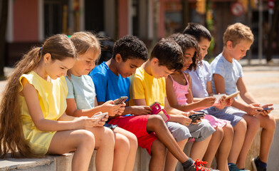 Group of positive children with mobile devices sitting at urban street