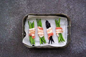 A baked appetizer, a bundle of green beans wrapped in bacon in a metal baking dish against a dark...