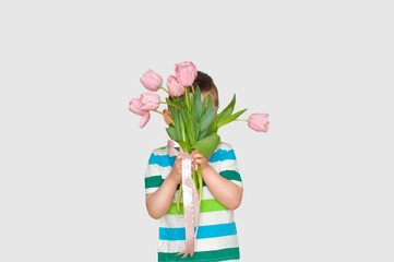 A cute little boy gentleman in a colored striped T-shirt holds a bouquet of pink tulips and hides behind it.