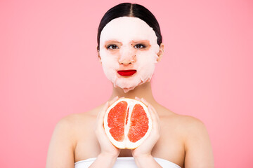 young asian woman in face mask holding half of ripe grapefruit isolated on pink