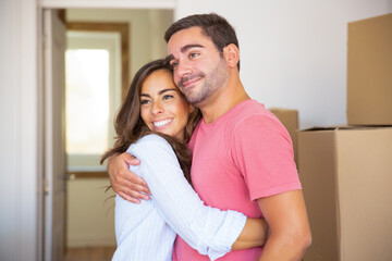 Joyful couple moving into new house, standing among carton boxes and hugging. Medium shot. New home or mortgage concept