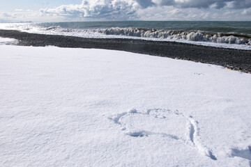 Obraz premium Heart shaped form on white snow with rocky beacj and sea view onthe background. Horizontal blank space image.