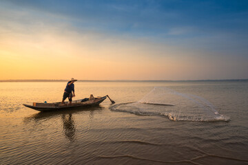 Asian fisherman on wooden boat throwing a net for catching freshwater fish in nature river in the early morning before sunrise