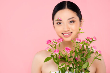 brunette asian woman with floral decoration on face holding tiny roses isolated on pink