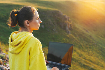 Young woman traveler freelancer in yellow hoodie with opened laptop on the beautiful view background