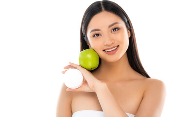 brunette asian woman holding green apple and cosmetic cream near face isolated on white