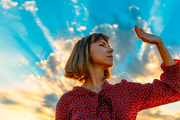 Young beautiful woman in a red dress against a blue sky with bright beams of the sun. The girl buries herself with one hand from the sun.