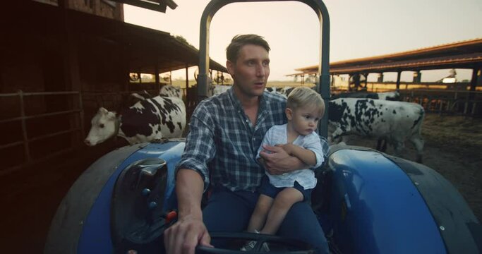 Authentic shot of happy farmer father is driving a tractor with his little son and showing a family dairy farm with ecologically grown cows. Concept: agriculture, farming, family, parenting, childhood