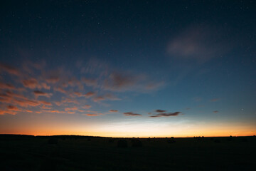 Night Starry Sky With Glowing Stars Above Countryside Landscape. Light Cloudiness Overcast Above Rural Field Meadow In Early Spring. Illumination Lights Of Town On Horizon