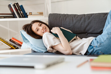 Image of cheerful beautiful girl reading book while lying on sofa