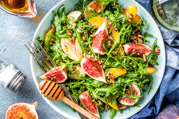 Autumn fig and arugula salad recipe. Whole vegan paleo fruit and vegetable fall salad idea. Homemade salad bowl with figs, arugula, peach and apple slices, nuts and honey. Blue concrete background cop