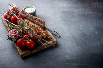 Shish kebab on skewers, from minced beef meat. Lula kebab, traditional Caucasian recipe. On black concrete background, with sauces, tomatoes and veggie salad top view copy space